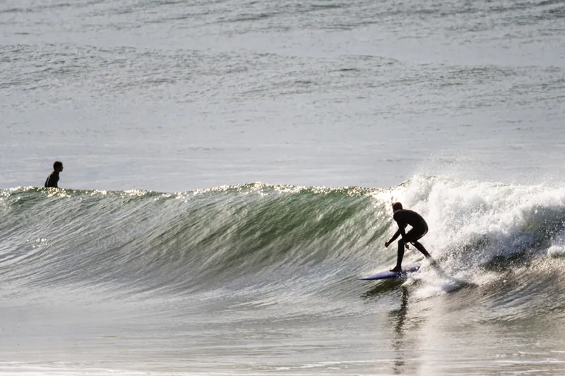 Surfista descendo a parede lisa de uma onda contra a luz, com outro surfista esperando a série ao fundo