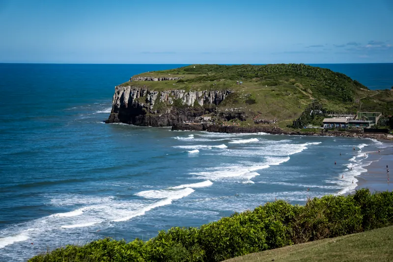 Vista do alto do promontório das Furnas avançando sobre o mar, com as ondas envolvendo a ponta e a Praia Grande ao fundo, em Torres (RS)
