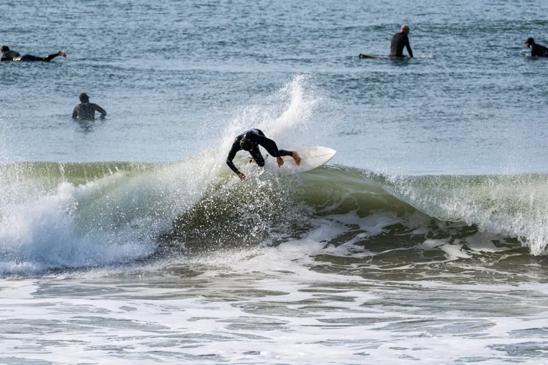 Surfista rasgando uma manobra na crista da onda, com leque de spray e outros surfistas ao fundo